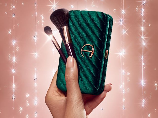 Close-up of a medium-tone model holding a makeup bag in a bottle green colour with the CT logo in the middle in golden with a contour blending brush and an eyeshadow brush. 