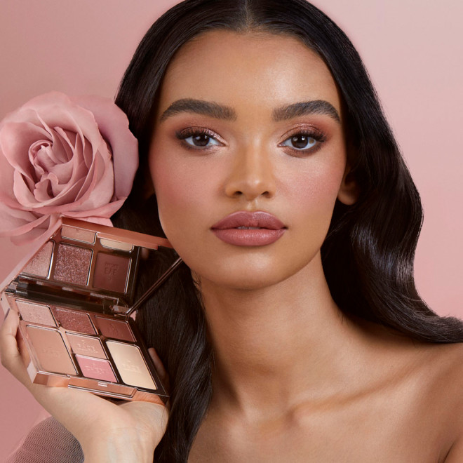 A medium-tone model with brown eyes wearing a soft-glam makeup look using the face palette she's holding that includes three eyeshadows in rose gold, reddish-pink, and dark brown shades, blush and highlighter in medium-pink and rose-gold, and contour powders.