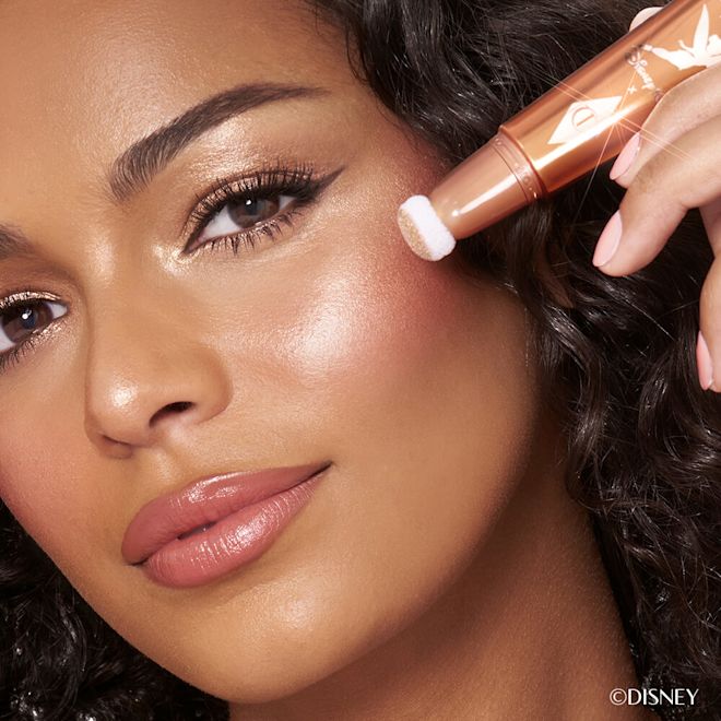 Model wearing Disney 100 x Charlotte Tilbury Beauty Light Wand in Spotlight Close up of brunette, tanned model wearing a glowy makeup look and holding Charlotte Tilbury's Disney liquid highlighter in rose gold shade.