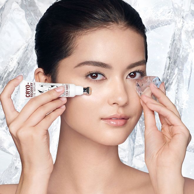 Cyro-Recovery Eye Serum on fair-tone model Fair-tone model with flawless, poreless, and glowy skin applying a white-coloured eye serum under her one eye and massaging an ice cube on the other side for a depuffing, cooling, and relaxing her skin.