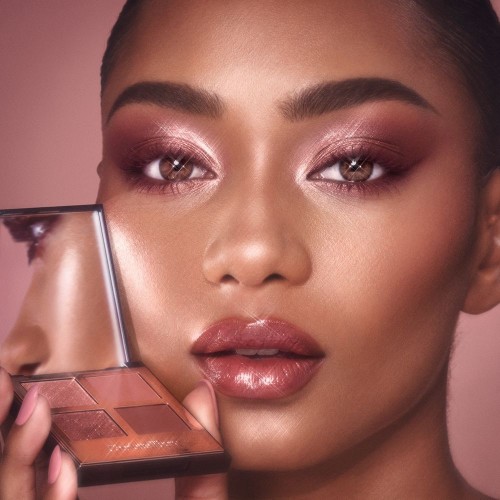 Deep-tone model with hazel eyes wearing berry pink lipstick with a gloss on top with eye makeup in shades of pearlescent rose gold, dusky rose, berry brown and rose-bud pink while holding up a quad eyeshadow palette used to create the eye look.
