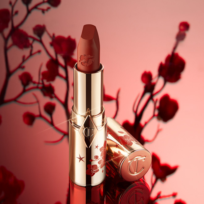 A matte burnt-orange red lipstick in gold-coloured packaging with cherry blossoms illustrated on the tube for the Lunar New Year, along with its lid next to it.