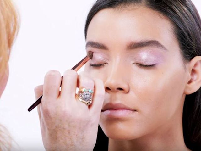 A light-tone brunette model with dewy skin getting a frosted lilac eyeshadow applied on her eye crease. 