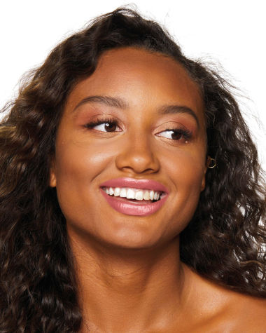Deep-tone model with brown eyes wearing a moisturising lipstick balm in a sheer nude pink berry shade with a high-shine finish.