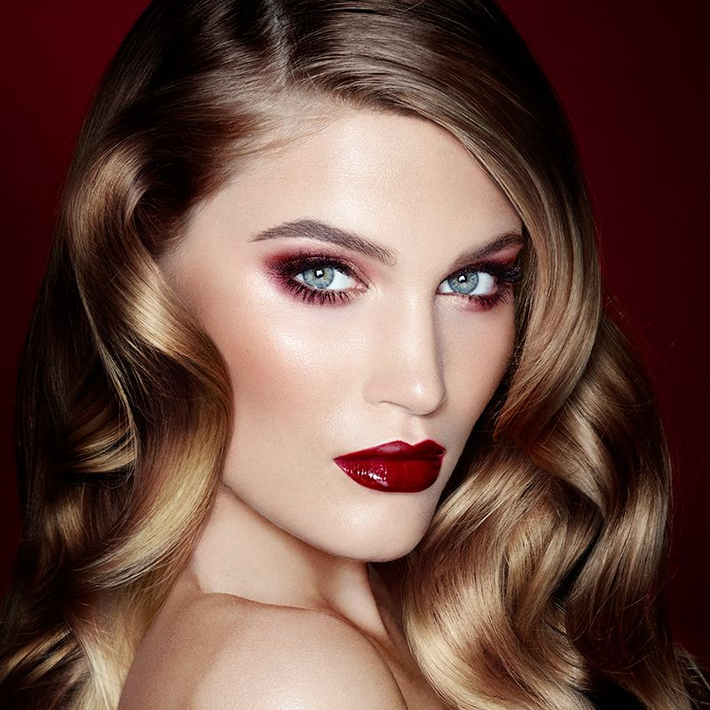 A fair-tone, blonde model with blue eyes wearing soft red, smokey makeup with a muted red blush, and vivid, crimson lipstick with a satin finish. 