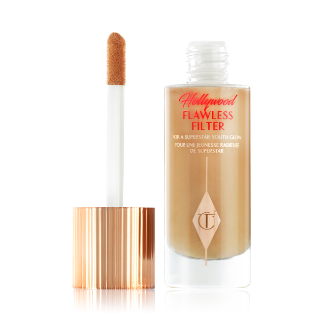 An open, luminous primer in a medium golden-beige shade in a glass bottle with its gold and white doe-foot applicator next to it.