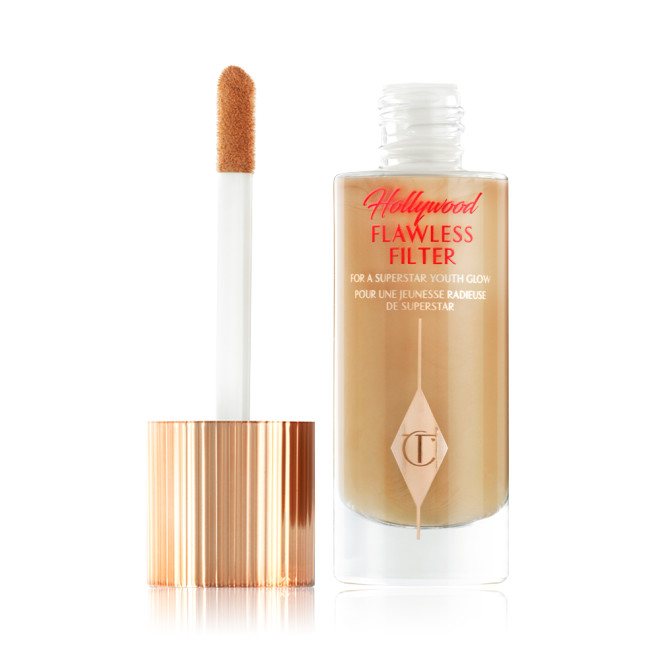 An open, luminous primer in a medium golden-beige shade in a glass bottle with its gold and white doe-foot applicator next to it.