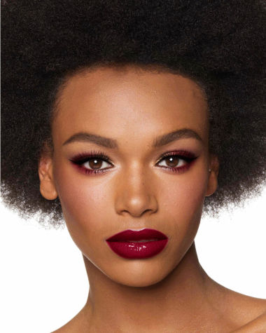 A deep-tone brunette model with reddish-plum eye makeup, glowy face base, and a vampy-red lipstick with a satin-finish. 