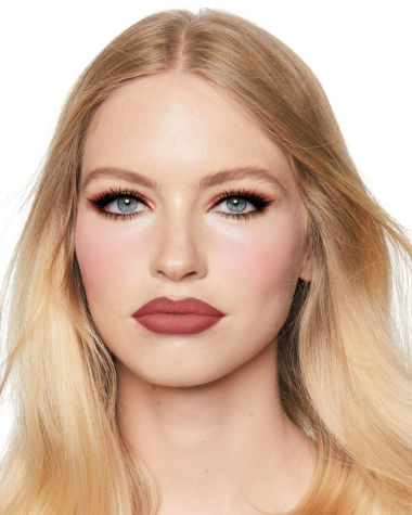 A fair-tone model with blue eyes wearing shimmery brown and gold eyeshadow with black kohl liner on her upper lid and lower waterline, a cool-toned glowy pink blush, and a berry-pink matte lipstick. 