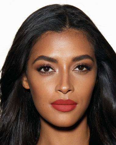 A deep-tone model with brown eyes wearing a muted, matte brick red lipstick. 