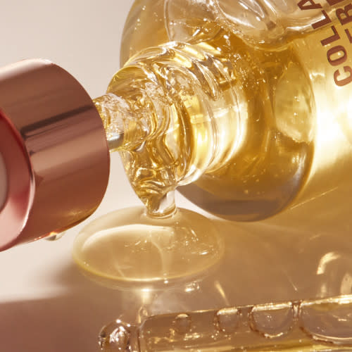 Close-up of luminous, light-gold-coloured facial oil pouring from a a glass bottle with a gold and white-coloured dropper lid.