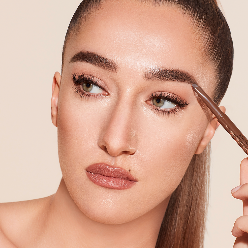 Light-tone model with green eyes filling in her brows with a brown-coloured eyebrow pencil.