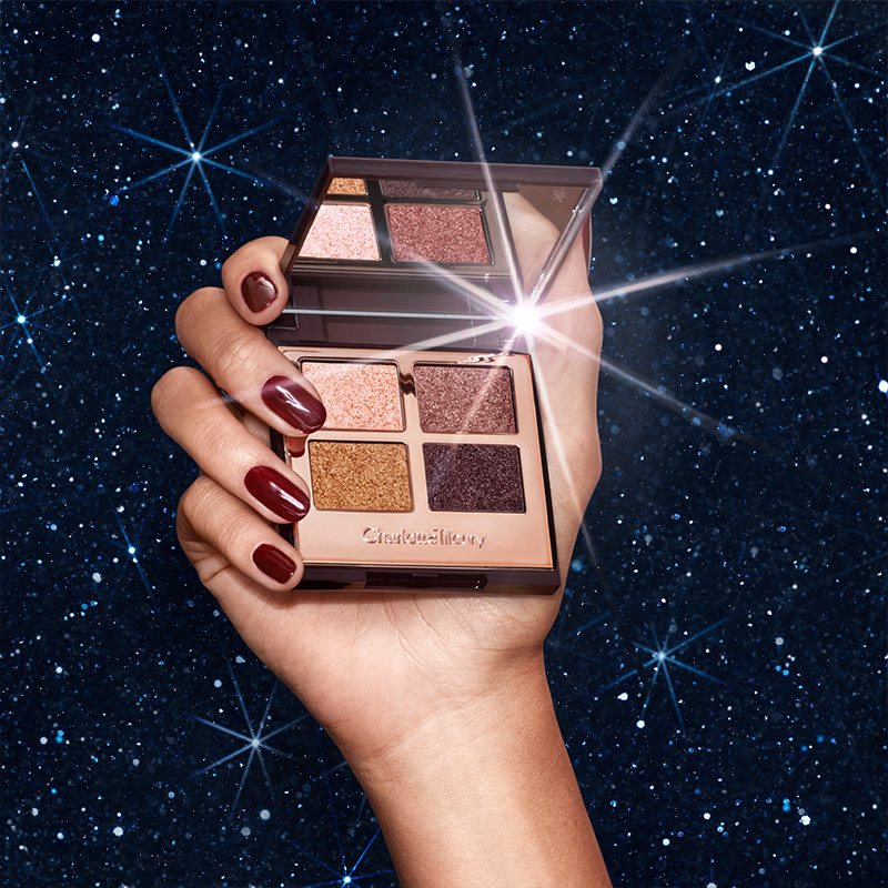 An open, mirrored-lid quad eyeshadow palette with shimmery eyeshadows in pink, gold, and purple held by a medium-tone model against a night-sky background. 