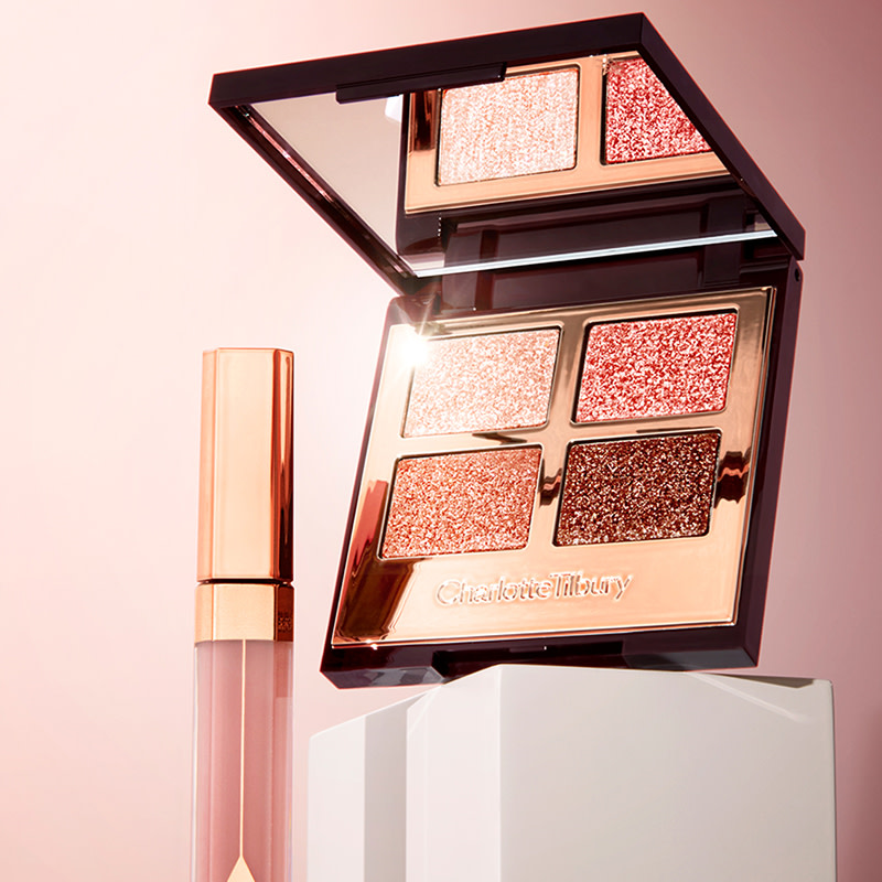 An open, mirrored-lid eyeshadow palette with shimmery eyeshadows in pink, copper, cream, and brown, and a lip gloss in nude pink with a golden-coloured lid. 