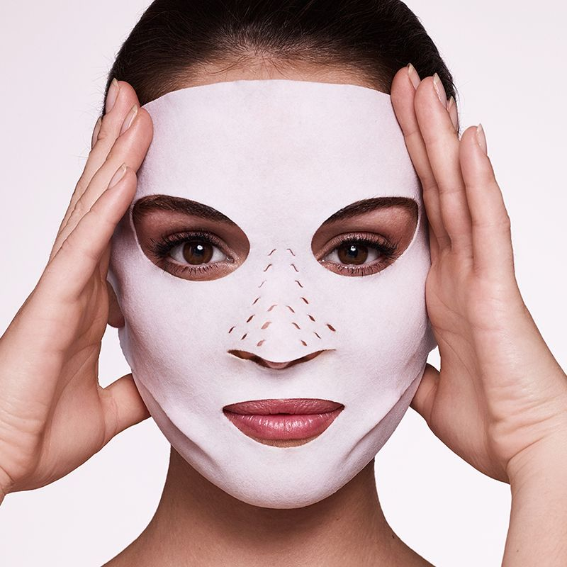 A medium-tone model wearing a dry sheet mask in textile fabric with ear holes and tiny holes on the nose area. 
