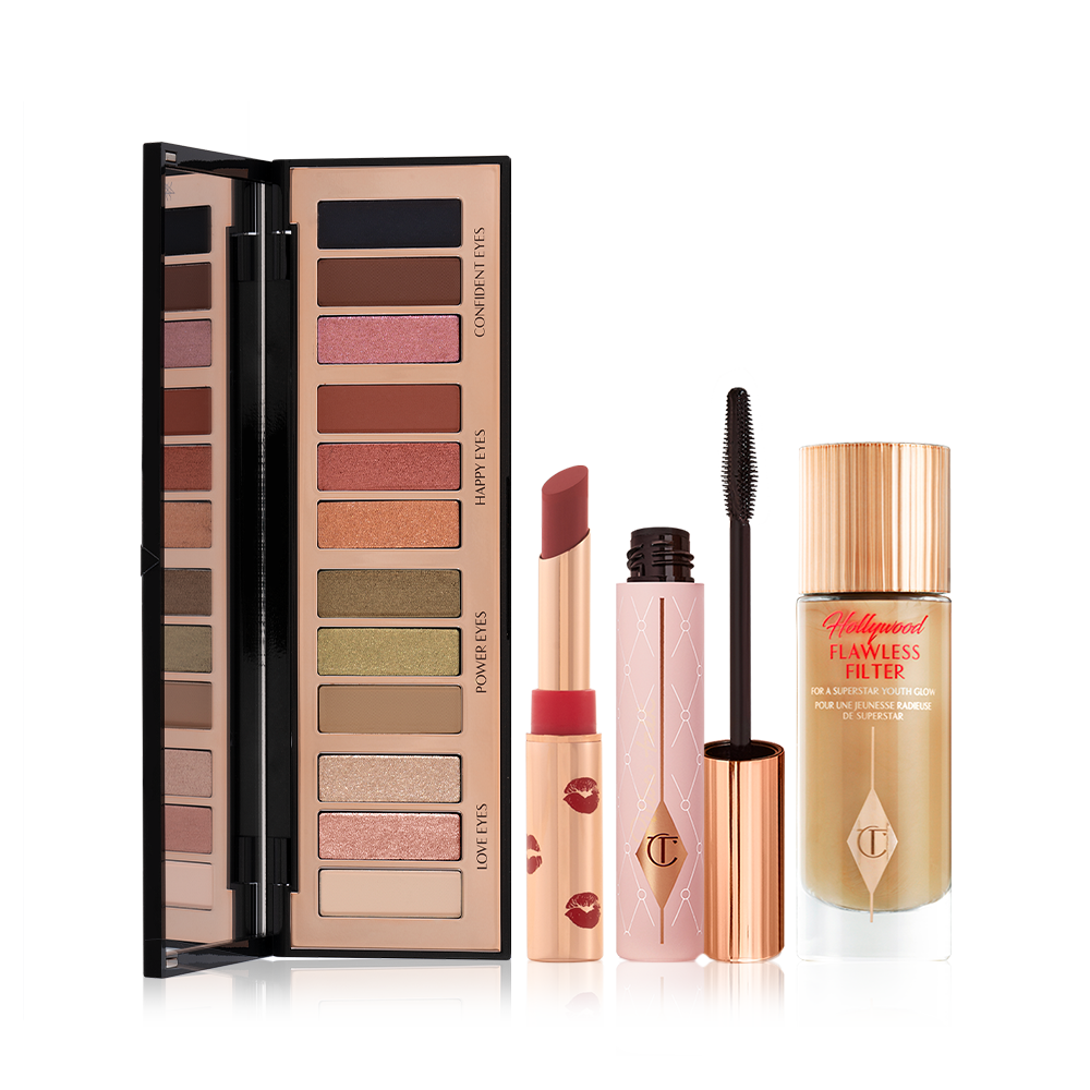 An open, mirrored-lid, 12-pan eyeshadow palette with earthy, matte and shimmery shades, an open matte lipstick in a dark red shade, mascara in nude pink tube with a gold-coloured lid, and glowy, skin-like primer illuminator in a glass bottle with a gold-coloured lid.