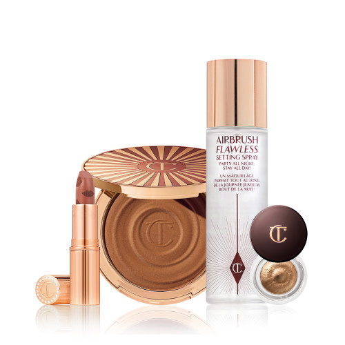 An open nude peach lipstick in a golden-coloured tube, cream bronzer compact with a mirrored lid, setting spray with a gold-coloured lid, and cream eyeshadow in a a glass pot with a dark brown coloured lid.