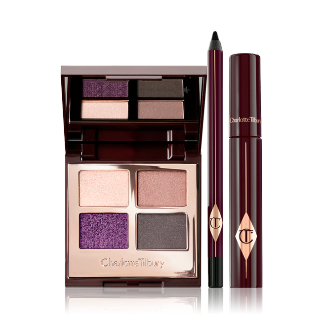 An open, mirrored-lid eyeshadow palette with eyeshadows in shades of purple, gold, and grey, an eyeliner pencil in black, and a black mascara in dark crimson packaging.