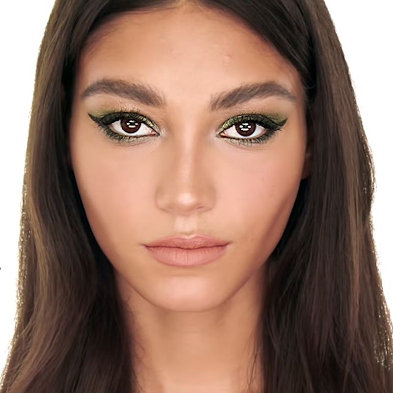 A light-tone brunette model with brown eyes wearing smokey green eyeshadow with shimmery green eyeliner paired with matte, nude peach lipstick. 