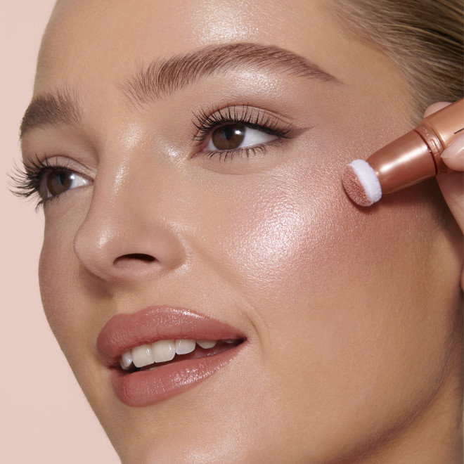 Medium-tone brunette model wearing glossy peach lipstick with shimmery gold and fawn eye makeup, and applying a glowy coral-peach blush from a liquid highlighter blush wand.