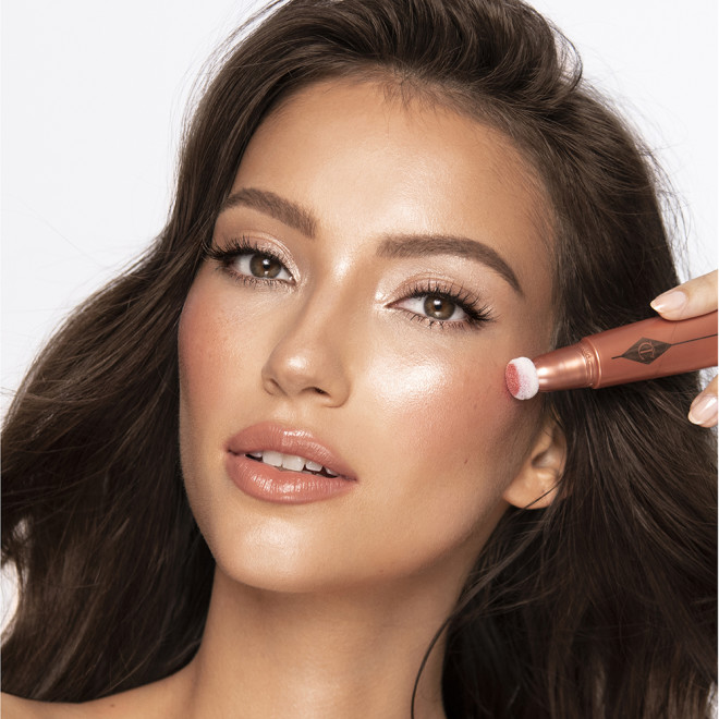 Medium-tone brunette model wearing glossy peach lipstick with shimmery gold and fawn eye makeup, and applying a glowy coral-peach blush from a liquid highlighter blush wand.