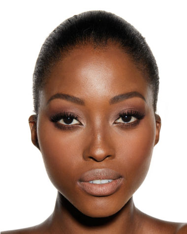 Deep-tone model with brown eyes wearing nude peach lipstick with soft coral blush, shimmery rose gold eyeshadow, and glow bronzer.