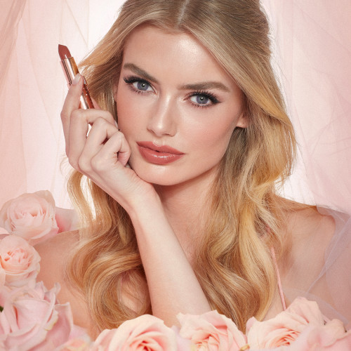 Fair-tone model with blue eyes wearing a universally flattering, peachy-nude lipstick with a satin-finish while holding the lipstick.