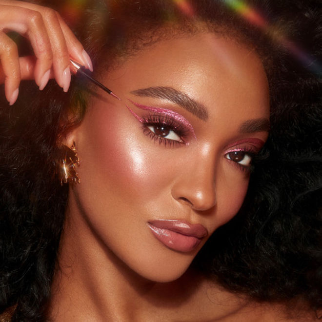 Pillow Talk Crystal Dimension Eyeliner on deep-tone model with brown eyes A deep-tone model with brown eyes applying a glittery vivid, rose pink eyeliner while wearing full-glam dewy and glowy pink makeup.