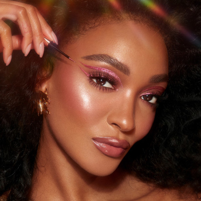 A deep-tone model with brown eyes applying a glittery vivid, rose pink eyeliner while wearing full-glam dewy and glowy pink makeup.