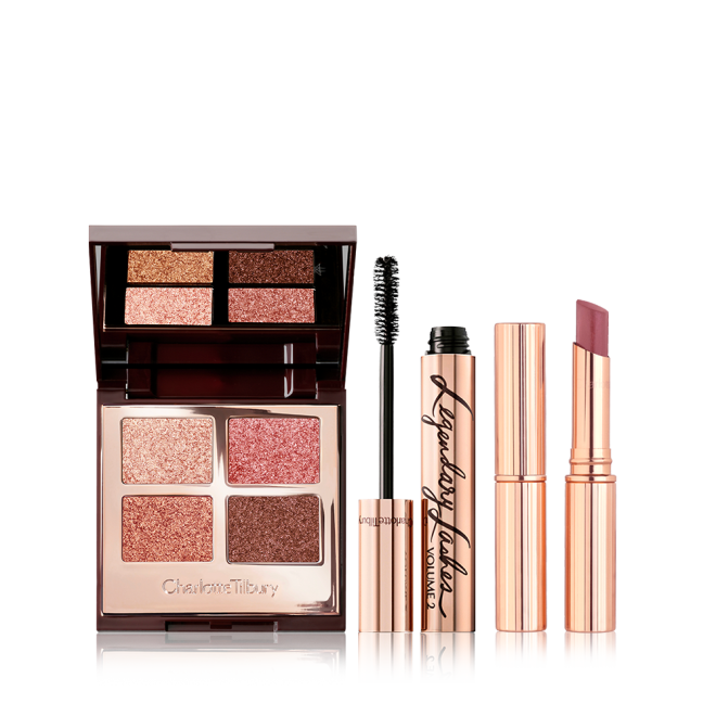 An open, mirrored-lid quad eyeshadow palette in shimmery pink and brown shades, a mascara in golden packaging with its applicator next to it, and a berry-pink lipstick. 