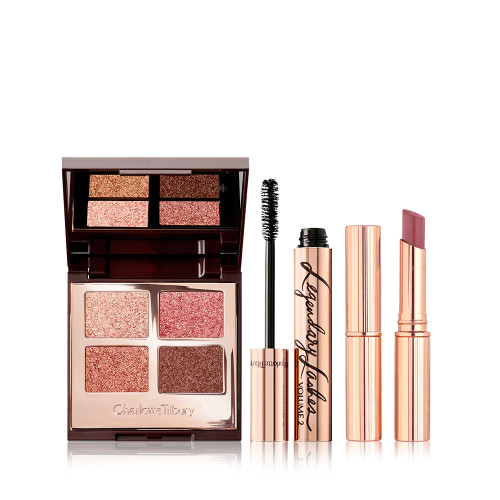 An open, mirrored-lid quad eyeshadow palette in shimmery pink and brown shades, a mascara in golden packaging with its applicator next to it, and a berry-pink lipstick. 