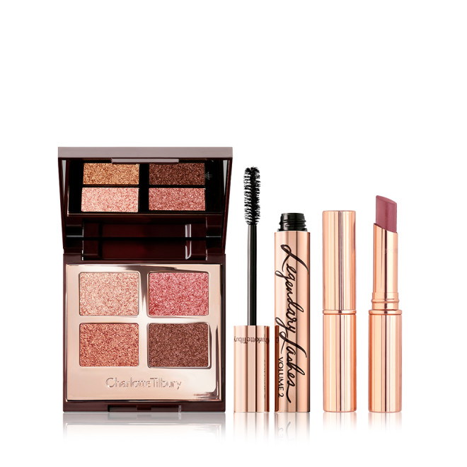 An open, mirrored-lid quad eyeshadow palette in shimmery pink and brown shades, a mascara in golden packaging with its applicator next to it, and a berry-pink lipstick. 