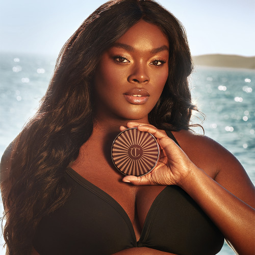 Deep-tone brunette model with glowy bronzed skin and soft-glam pink makeup holding a closed cream bronzer compact.