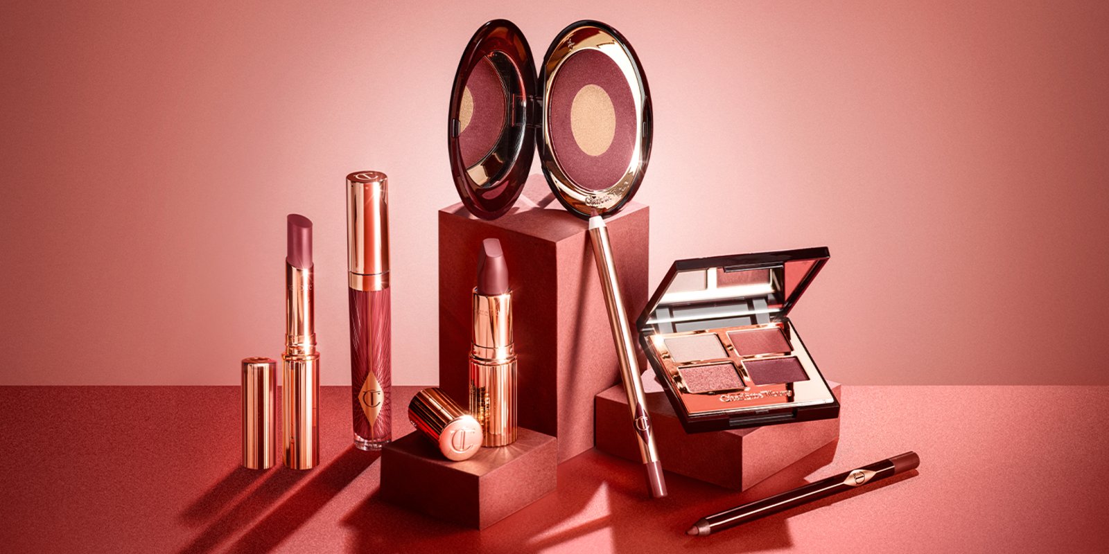 A collection of different makeup products that include lip gloss, lipstick, lip liner pencils, eyeliner pencil, lipstick lip balm, two-tone blush, and quad eyeshadow palette in berry pink colours. 