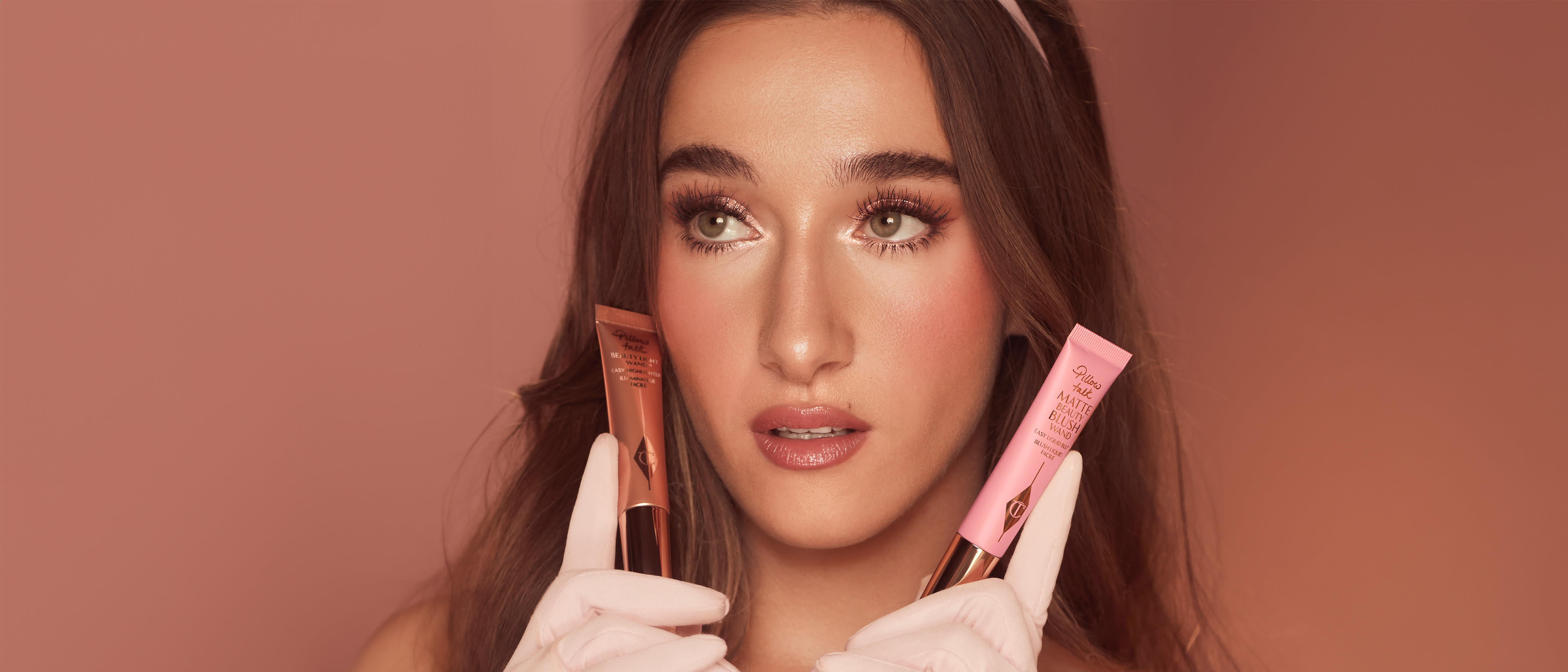 Brunette model wearing pale pink gloves and pretty pink wedding makeup holding Charlotte Tilbury highlighter and blush wands