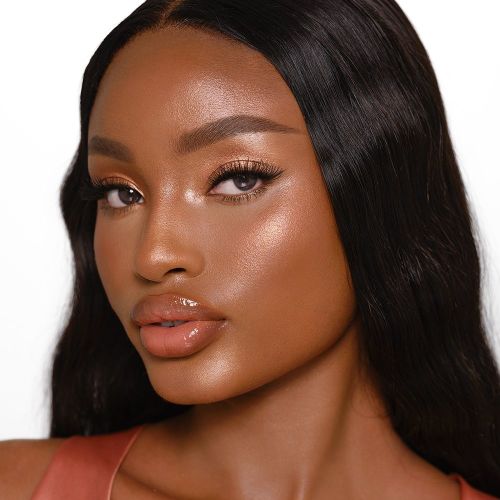 Deep-tone model with brown eyes wearing a soft rose gold highlighter with nude brown lip gloss and jet black eyeliner.