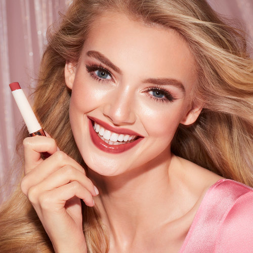 Fair-tone model with blue eyes wearing a moisturising lipstick balm in a soft brown shade with a high-shine finish while holding the lip balm.