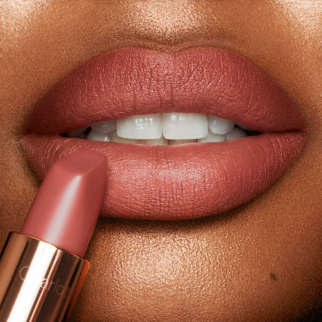 Close-up of a deep-tone model applying a warm, peachy-nude matte lipstick.