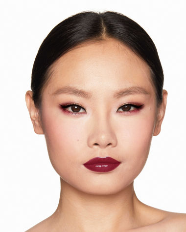 A fair-tone model with brown eyes wearing reddish-plum eye makeup, glowy face base, and a vampy-red lipstick with a satin-finish. 