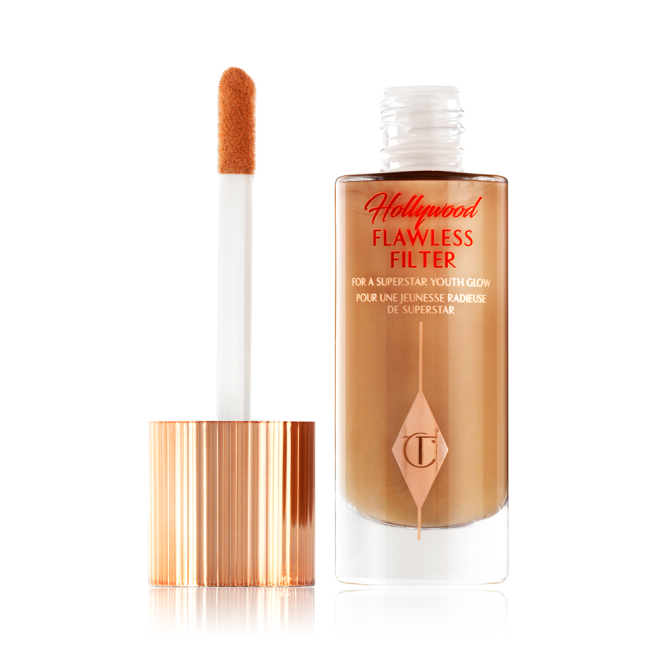 An open, luminous primer in a dark golden-beige shade in a glass bottle with its gold and white doe-foot applicator next to it.