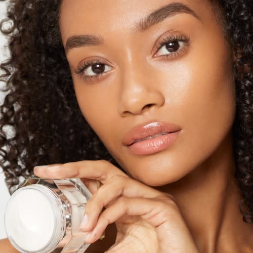 A deep-tone brunette model with glowy, flawless skin wearing a glowy and nourishing face cream and holding the face cream.