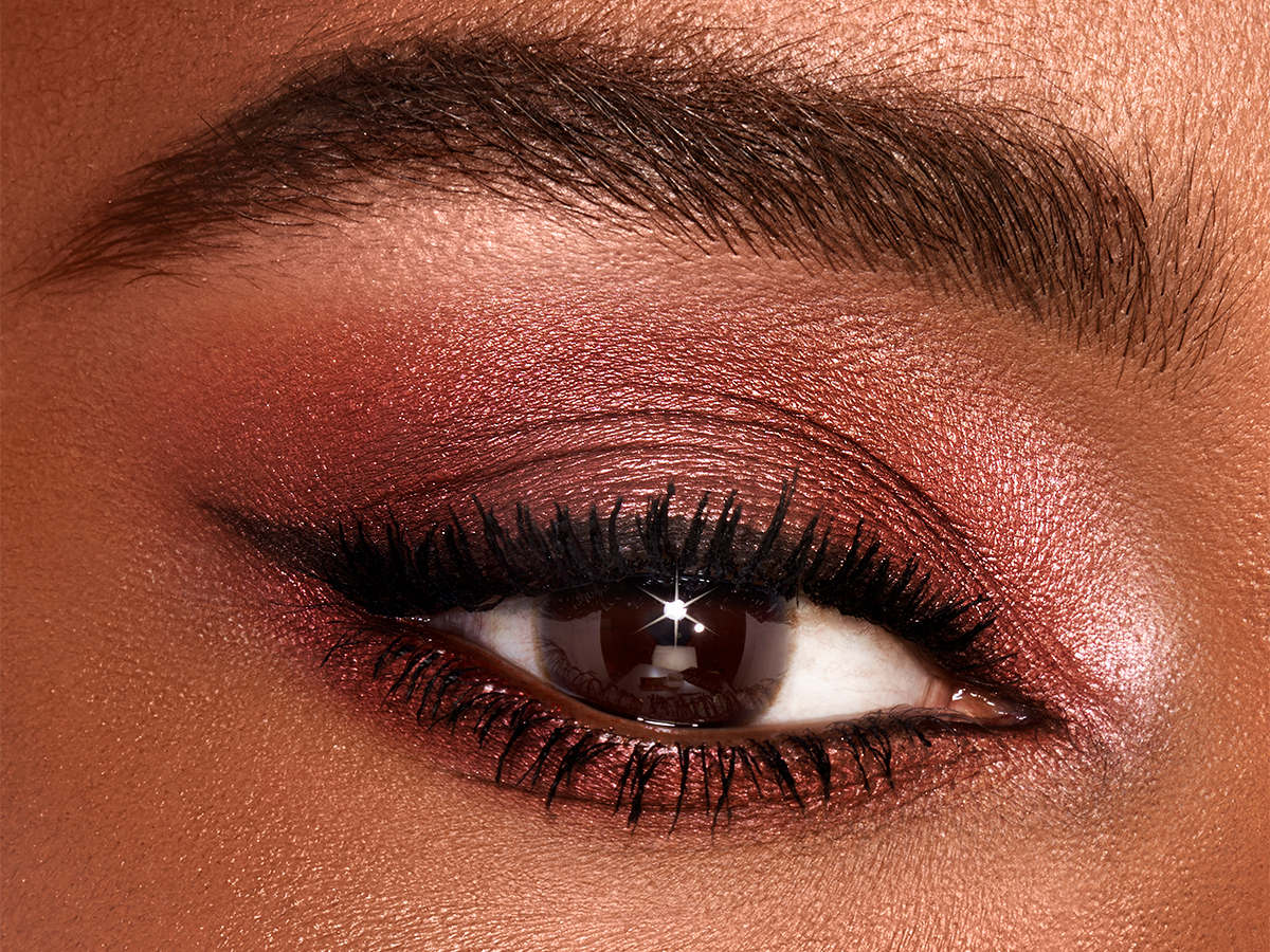 Single-eye close-up of a deep-tone model with brown eyes wearing shimmery red and rose-gold eyeshadow. 