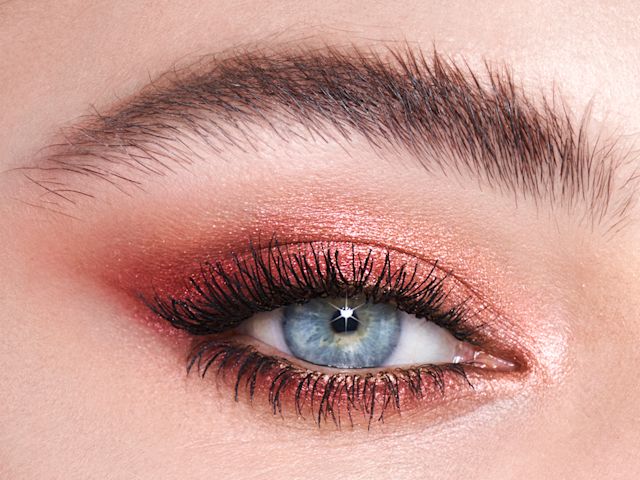 Single-eye close-up of a fair-tone model with blue eyes wearing shimmery red and rose-gold eyeshadow. 
