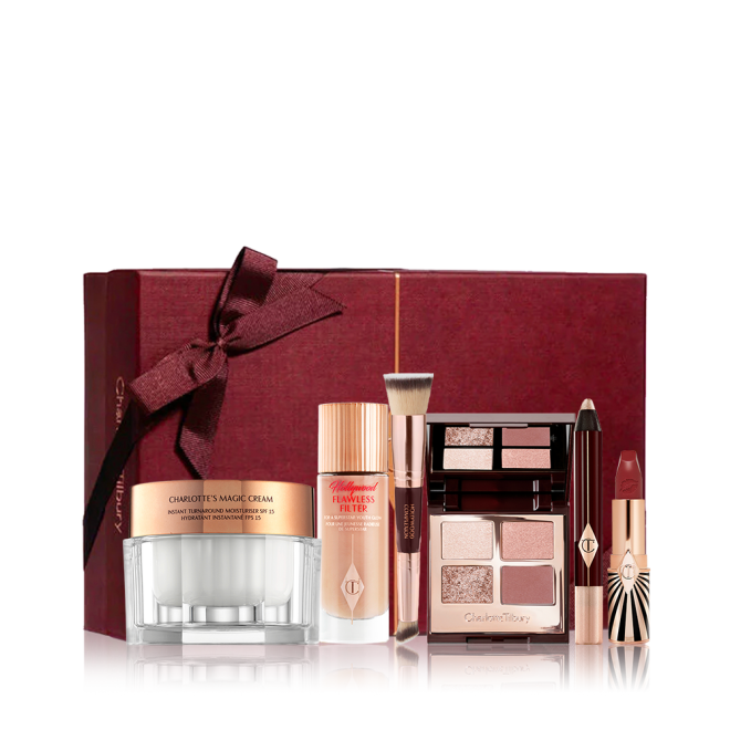 A pearly-white face cream in a glass jar with a glow-boosting luminous primer in a glass bottle, a double-sided face blending brush, ope, quad eyeshadow palette with matte and shimmery nude pink and brown shades, chubby eyeshadow stick in champagne, and an open lipstick in a crimson shade with a crimson coloured gift box in the back with a bow on it. 