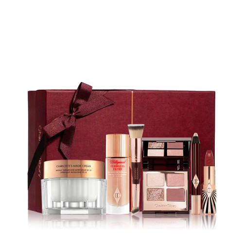 A pearly-white face cream in a glass jar with a glow-boosting luminous primer in a glass bottle, a double-sided face blending brush, ope, quad eyeshadow palette with matte and shimmery nude pink and brown shades, chubby eyeshadow stick in champagne, and an open lipstick in a crimson shade with a crimson coloured gift box in the back with a bow on it. 