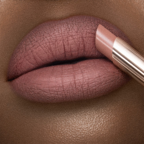 Lips close-up of a deep-tone model applying a warm peach-nude lipstick with a matte finish. 