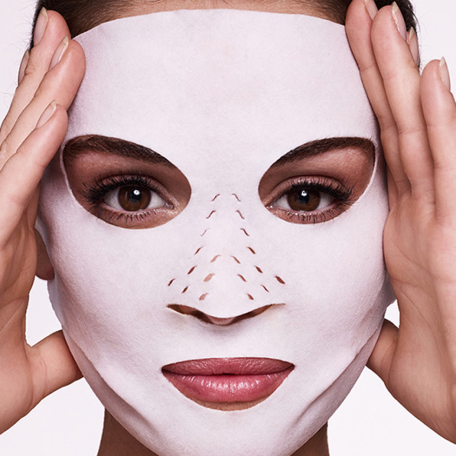 Close-up of a light-tone, brunette model wearing a sheet mask made of textile fabric with eyes, lips, and nose openings. 