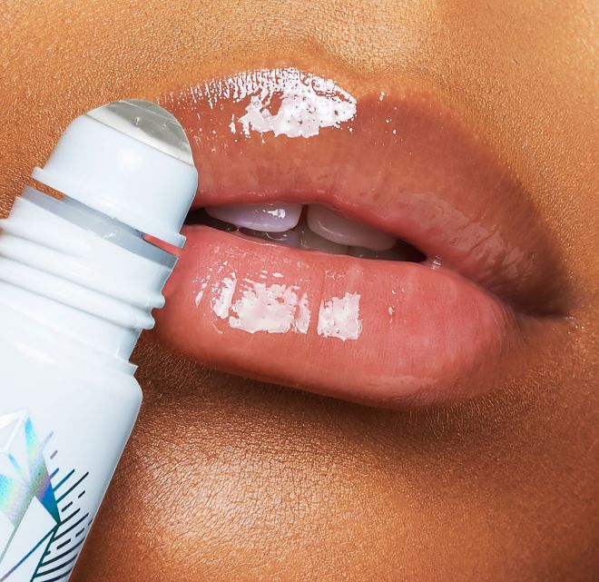 Close-up of a deep-tone model applying a nourishing, clear lip oil with a crystal roller ball applicator. 