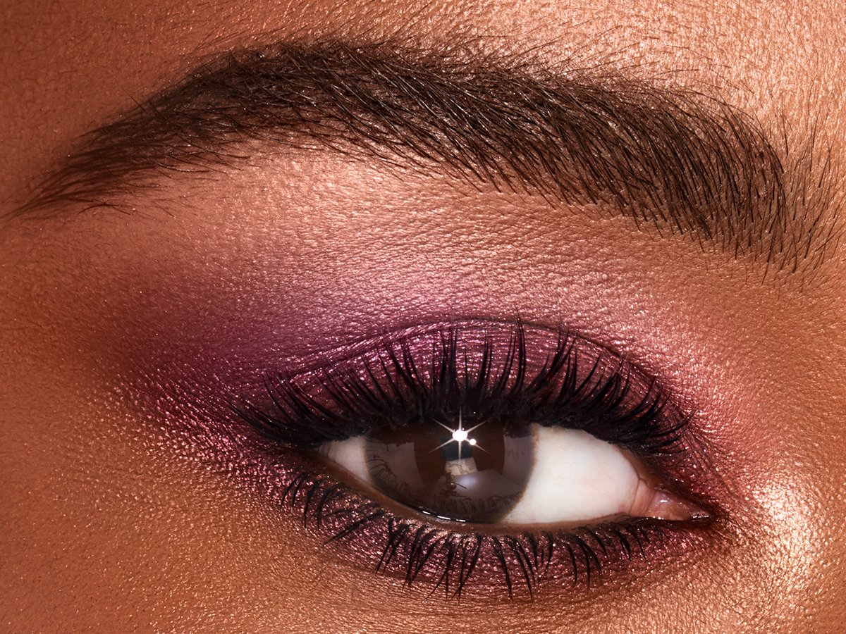 Single-eye close-up of a deep-tone model with brown eyes wearing shimmery rose gold and purple eyeshadow. 