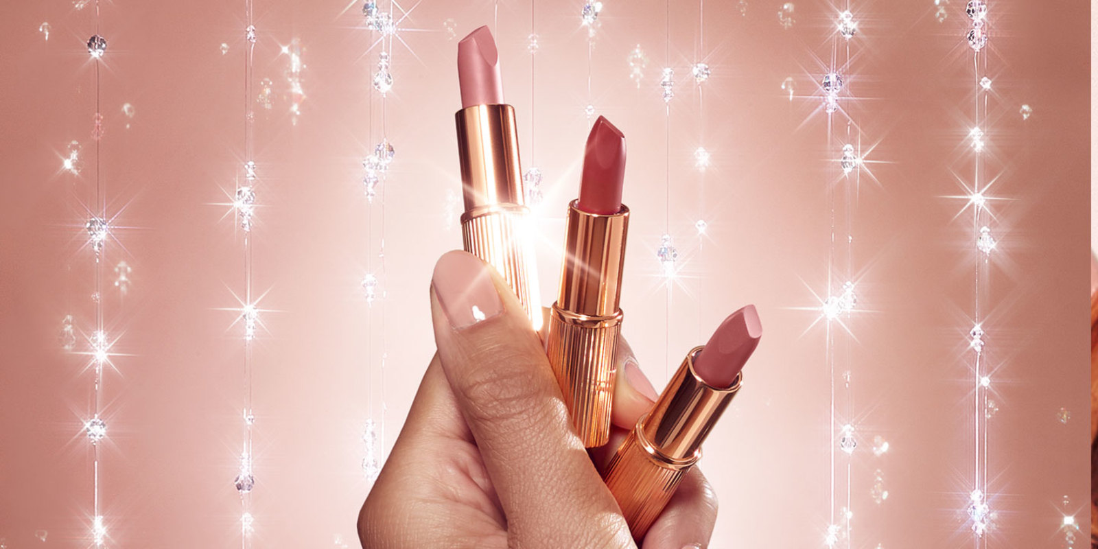 Close-up of a medium-tone model's hand holding three mini open lipsticks in nude pink, brown-pink, and statement red. 